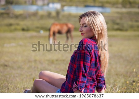 Young beautiful woman in checkered shirt and jeans shorts with blond hair in farm on the background of red horse.