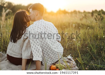 Stylish beautiful couple kissing on blanket in warm sunny light among grass in summer meadow, enjoying sunset. Vacation and picnic. Young boho family embracing, atmospheric beautiful moment