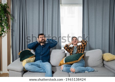 Young couple is sitting on a sofa in their apartment looking up and holding their hands to plug their ears as a neighbor upstairs is having a party and playing loud music or renovating the apartment  Royalty-Free Stock Photo #1965138424