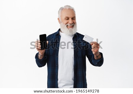 Smiling mature man showing smartphone blank screen, looking satisfied and happy at credit card of copyspace bank, standing against white background