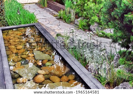 Decorative Above Ground Pond in Japanese Style in Back Yard Garden And Stone Footpath. Modern Backyard Garden Landscaping with Designed Elements. Family Rest Area at Small Designed Pond In Garden.