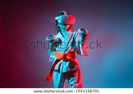Studio Portrait of young caucasian girl in karate uniform playing video games with virtual reality headset or practicing online with a trainer on blue and red background. Future technology concept. VR