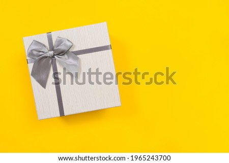 Close white box with lid decorated with beautiful silver ribbon bow for gift packaging, yellow background, top view, copy space for advertising text. Holiday stuff concept.