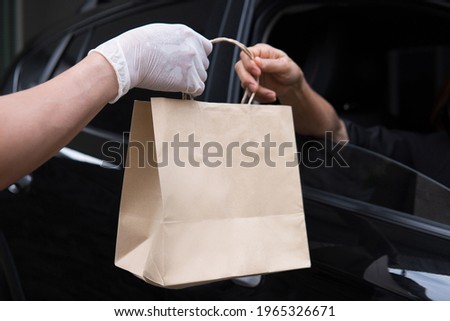 Closeup food delivery man hand wear glove passing brown paper bag to woman customer driving thru pickup out of black car window meeting social distancing requirements and supporting small businesses Royalty-Free Stock Photo #1965326671