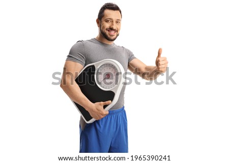 Young man in sport clothes holding a measuring scale and gesturing  thumb up sign isolated on white background Royalty-Free Stock Photo #1965390241