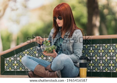 Red-haired woman with sunglasses sitting on a park bench eating healthy food on a sunny day. Full shoot.