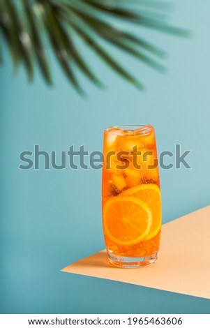 Cold cocktail with orange juice and ice on a light blue background with a leaf of a palm tree. Tall glass on a beige background with condensation. Chilled delicious summer drink.