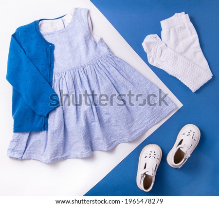Flat lay composition in blue and white color with fashion kid's clothes. There are blue dress, dark blue cardigan, white pantyhose and white sandals on the photography.  