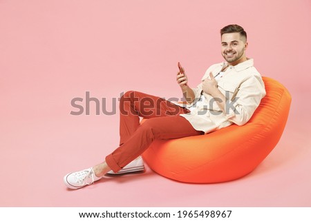 Full length young happy fun man in jacket white t-shirt sitting in bean bag chair using mobile cell phone chat online in social network show thumb up gesture isolated on pastel pink background studio. Royalty-Free Stock Photo #1965498967
