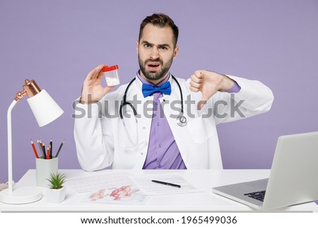 Male doctor man in white medical suit sits at desk work in hospital clinic office hold medication tablets aspirin pills in bottle show thumb down gesture isolated on violet background studio portrait.