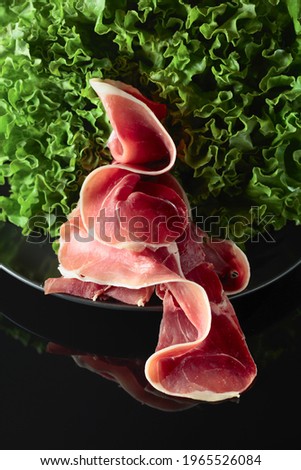 Prosciutto and fresh lettuce salad on a black reflective background.