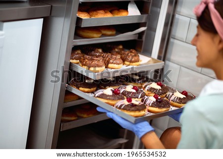 A young small business female owner fills the closet with delicious handmade donuts in a pleasant atmosphere in a candy workshop. Pastry, dessert, sweet, making Royalty-Free Stock Photo #1965534532