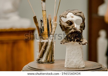 Focus on a Small bust of man, in white plaster, undergoing restoration next to a glass jar with art tools Royalty-Free Stock Photo #1965560434