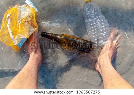 Top view man's feet on the beach surrounded by empty bottles, plastic and other garbage.