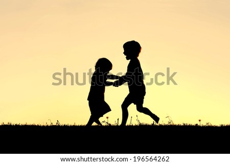 A silhouette of two happy little children, a young boy and his baby brother, holding hands and dancing and playing in front of a sunset in the sky.