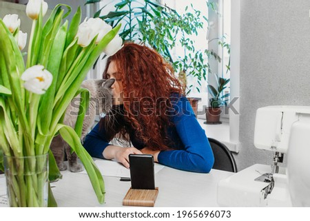 A middle-aged red-haired European woman kisses a British cat on the forehead. In front of her phone flowers tulips sewing machine cat prevents the hostess blogger to broadcast Home hobbies or training