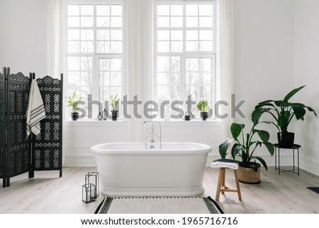 Home decor ideas in contemporary bathroom design. Empty freestanding bathtub against large windows, wooden folding screen and green house plants on floor. Concept of classic bath in modern apartment Royalty-Free Stock Photo #1965716716