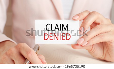 Woman writes in a notebook with a silver pen and hand hold card with text: claim denied. Pink background, front view. Business and education concept.