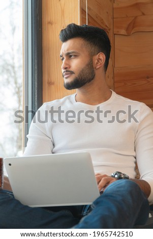 Young man wearing a white shirt and jeans sitting next to the window with his laptop. Looking outside through the window in a cozy room. Winter season in the city of Zakopane, Poland. 