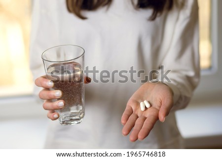 A woman holds a glass of water and two pills in her hands. Young woman treats illness with pills. Headache or stomach problems and treatment with medication. Vitamins as a supplement to nutrition Royalty-Free Stock Photo #1965746818