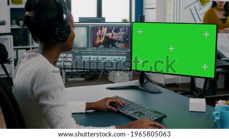 African woman videographer with headset processing film montage working at greenscreen, chroma key isolated display of computer. Video editor editing movie in post production software in agency office