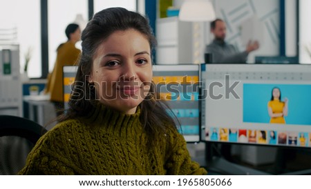 Photo retoucher sitting in creative workplace looking at camera smiling working in start up media agency. Woman editor retouching client photos on PC with two displays on editing app, software