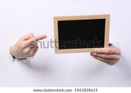 Female hands holding blank chalk board through torn hole white paper. Copy space