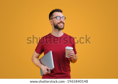 Portrait of confident young smiling bearded man in casual red t-shity and trendy spectacles holding laptop and paper cup of coffee, isolated on yellow background. Royalty-Free Stock Photo #1965840148