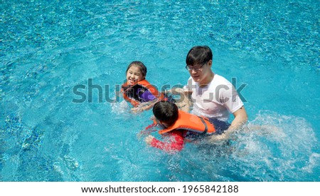 An Asian father teaches his son and daughter to swim in the pool. The older sister and younger brother wearing orange life jacket.
