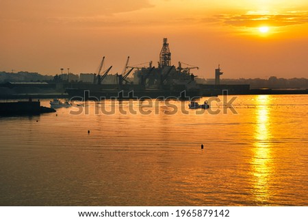 scene of fishing boats entering port at dawn with harbor cranes, an oil platform and the city skyline in the background