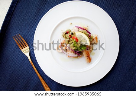 Baked Cod fish with green sauce (salsa verde) with lemon, red onion, coriander, red chili, pickles and capers. Basque cuisine. On white plate. On the blue background. Close-up.