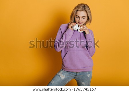 Portrait of a young sporty woman in blue jeans with abrasions, purple hoodie, sneakers and white wireless headphones in studio on a yellow background. Joy, happiness, cute blonde girl listen music