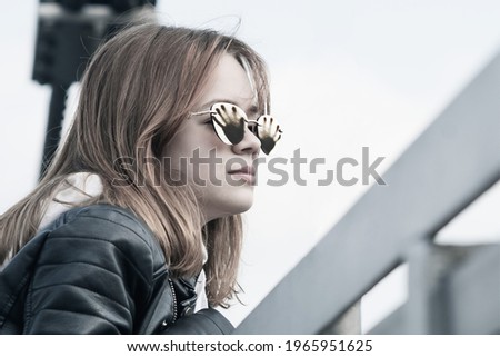 A young girl wearing glasses and a leather jacket walks in the park near the suspension bridge