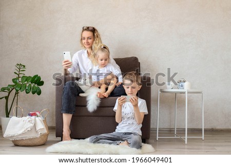 Mother and children are sitting in the living room on a chair with phones in their hands. Mom writes a message, makes purchases online, the child plays on the phone, watches cartoons. Family leisure. 