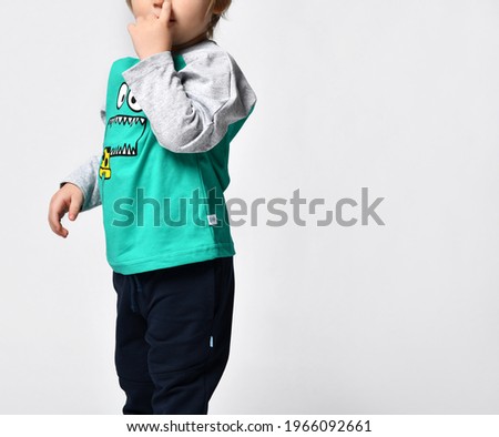 Cropped photo of a child boy in green long-sleeved casual clothes, falling asleep with his finger in his mouth, against a gray wall background. Happy childhood and fashionable children's clothing