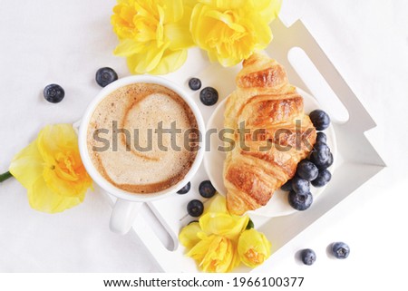 Morning breakfast in bed with yellow daffodils. Wooden white tray with a cup of coffee croissant  and blueberries. Morning at home. Relaxing atmosphere. Breakfast in the bed on the white  bedsheet.   