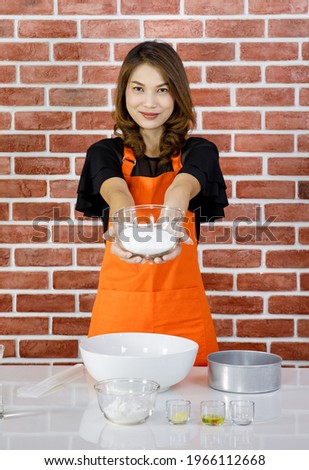 Asian young pastry chef lady in orange apron standing smiling look at camera showing glass bowl of white flour in hand when teaching baking and cooking in front red brick wall kitchen background. Royalty-Free Stock Photo #1966112668