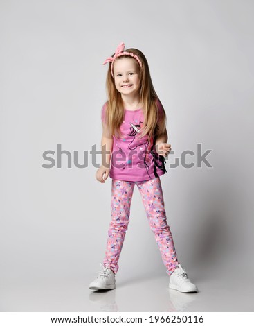 Active playful blonde kid girl in summer clothing colorful pants and t-shirt stands laughs and giggles over light background