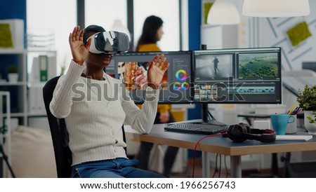 African video editor experiencing VR goggles, gesturing, editing video film montage working with footage and sound on computer with two displays. Videographer processing movie in creative agency