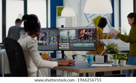Black videographer putting on headset editing movie using post production software working in creative start up agency office. African editor processing video footage on computer with dual displays