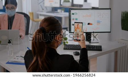 Executive business woman wearing protective face mask using phone for online videocall conference with remotely team. Coworkers working in background respecting social distance