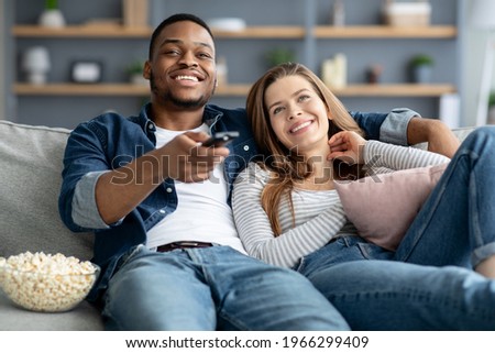 Domestic Pastime. Happy Mixed Race Couple With Popcorn Watching TV At Home, Loving Interracial Spouses Resting Cn On Comfortable Sofa In Living Room, Man Switching Channels With Remote Controller