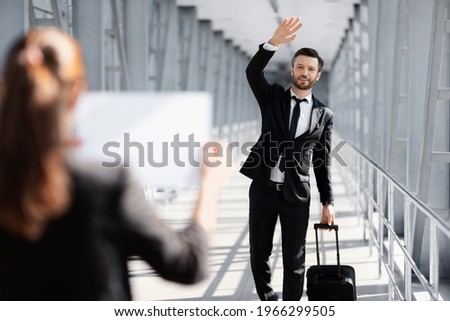 Cheerful businessman waving to assistance with placard, meeting in airport. Handsome middle-aged entrepreneur with luggage arrived for business meeting, greeting business partner or representative Royalty-Free Stock Photo #1966299505