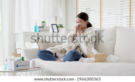 Sick young asian woman sitting under the blanket on sofa and sneeze with tissue paper at home. Female blowing nose, coughing or sneezing in tissue at home, suffering from flu. Cold and fever concept Royalty-Free Stock Photo #1966300363