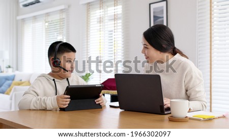 Young adult asian mother and kid stay home work and learning online remotely from computer tablet in self isolate from lockdown coronavirus covid19 social distance. Homeschool boy with parent. Royalty-Free Stock Photo #1966302097