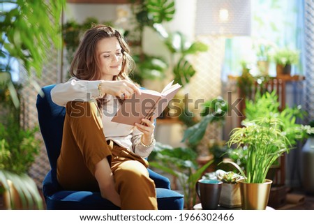 Green Home. relaxed trendy middle aged woman with long wavy hair with book in green pants and grey blouse in the modern living room in sunny day. Royalty-Free Stock Photo #1966302466