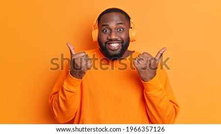 Positive black man with thick beard makes like gesture keeps thumbs up enjoys favorite track list in headphones wears casual long sleeved jumper isolated over vivid orange background. Happy meloman