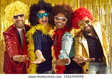 Pop music band singing at night club. Young people disguised in fancy feather boas and funny silly curly wigs dancing together. Happy multiethnic men and women having fun at Halloween disco party Royalty-Free Stock Photo #1966394098