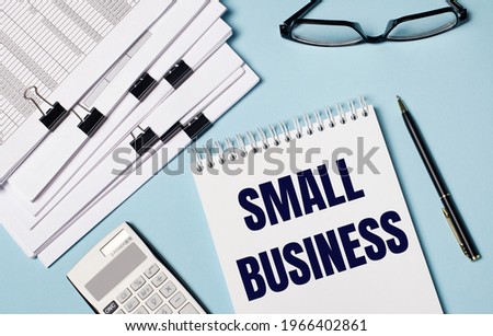 On a light blue background there are documents, glasses, a calculator, a pen and a notebook with the text SMALL BUSINESS. Close-up of the workplace. Business concept