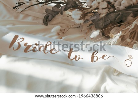 Bride to be golden and white satin sash ribbon accessory for a bachelorette party and boho neutral dried flowers on the background Royalty-Free Stock Photo #1966436806